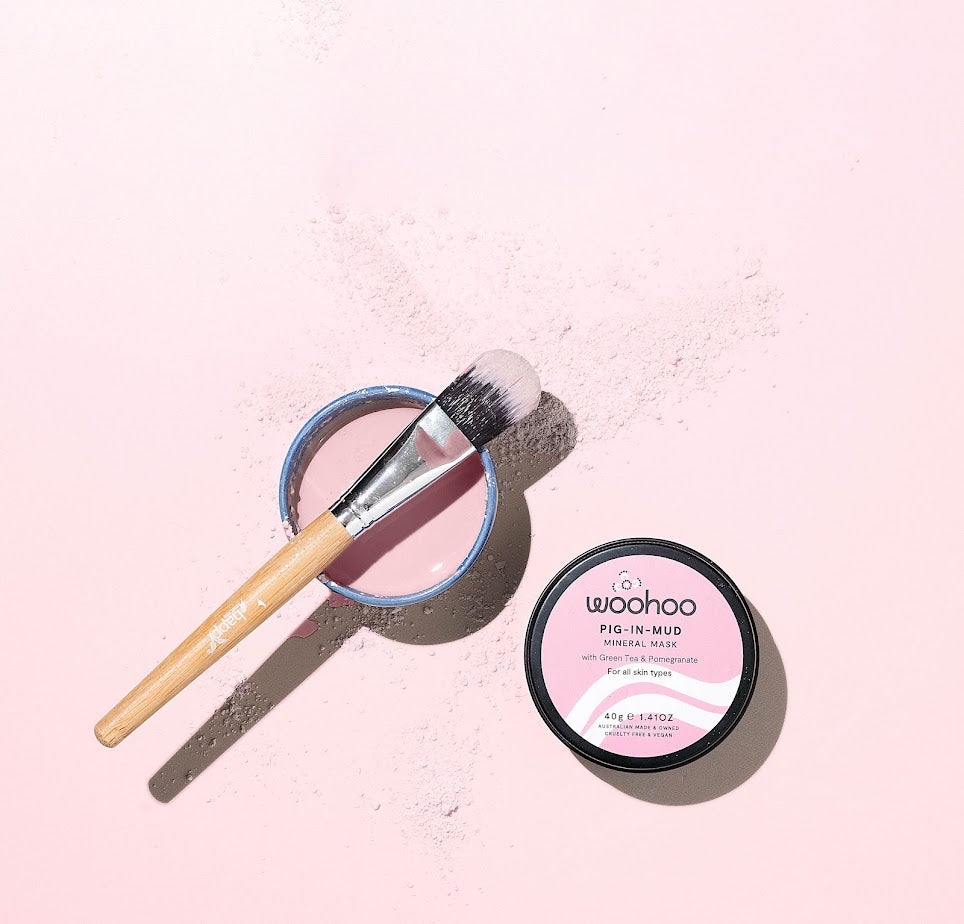 Image of the Woohoo Pig-In-Mud Mineral Mask product and mixed in a small bowl and a brush on it with some of the dry product around on a pink background