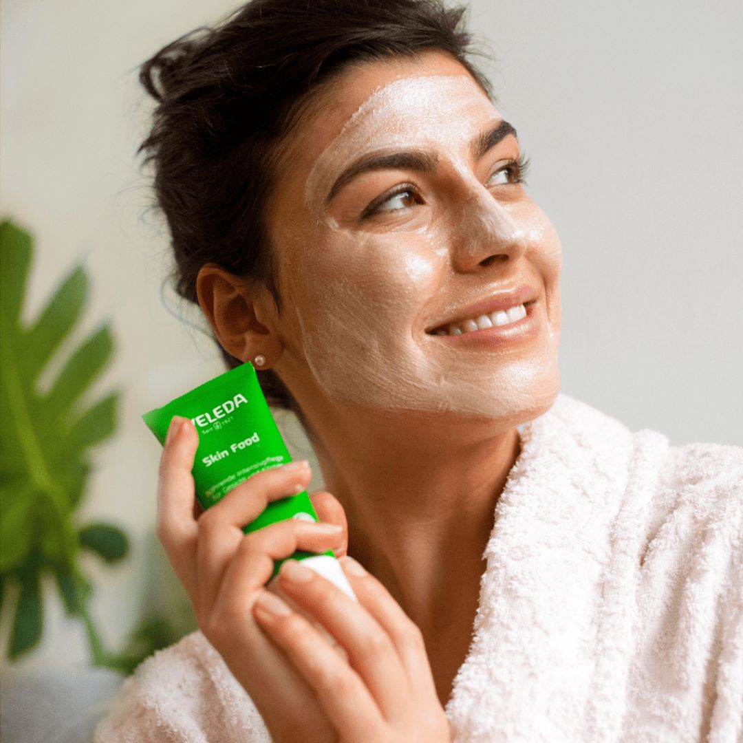 Woman with brown hair holding a tube of Weleda Skin Food and demonstrating Weleda Skin Food as a face mask