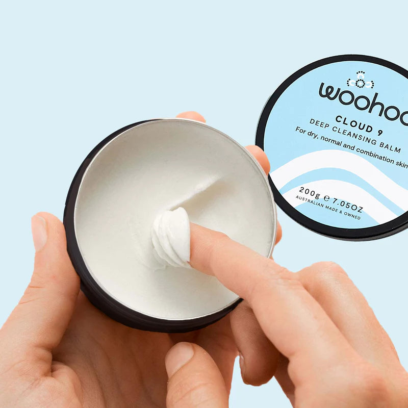 Image of the Woohoo Cloud 9 Deep Cleansing Balm tin opened and held by one hand with the finger from the other hand dipping into the product on a light blue background 