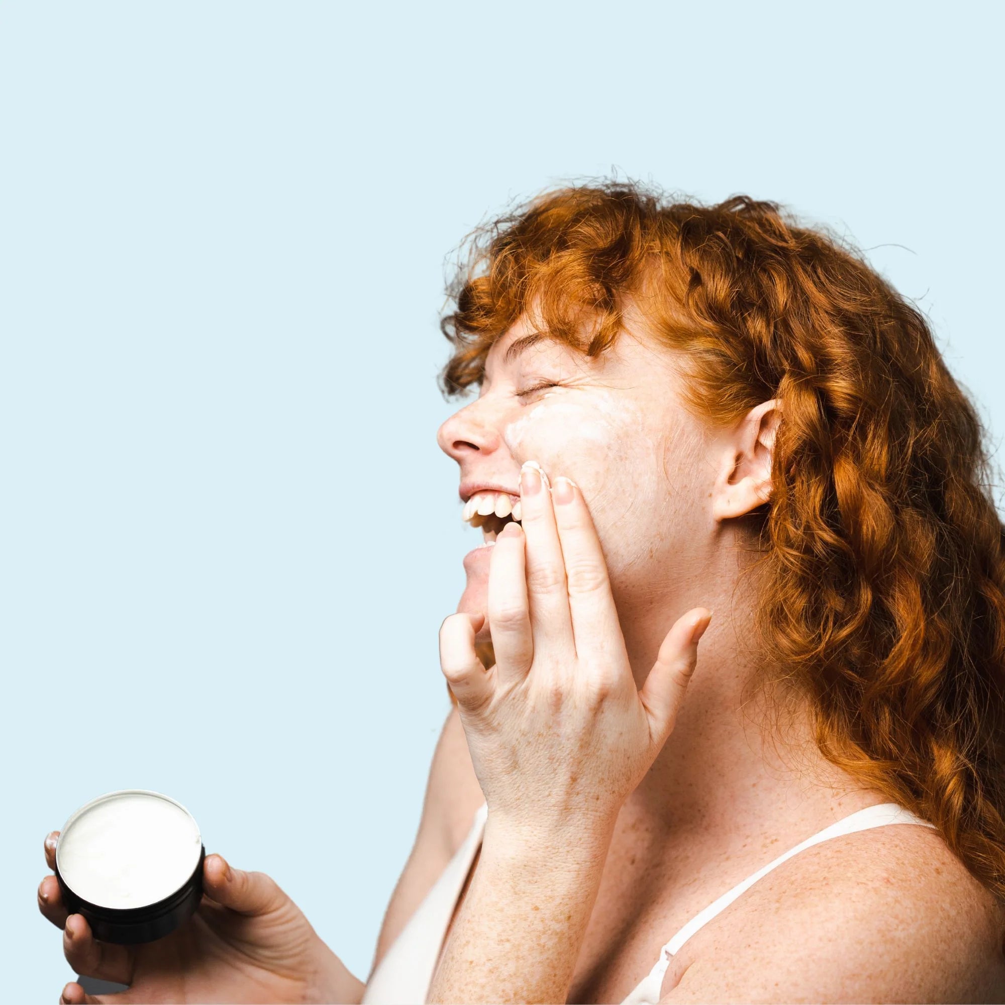 Image of a redheaded woman in a white singlet holding the opened Woohoo Cloud 9 Deep Cleansing Balm Tin and applying the product on her cheek from the side profile.