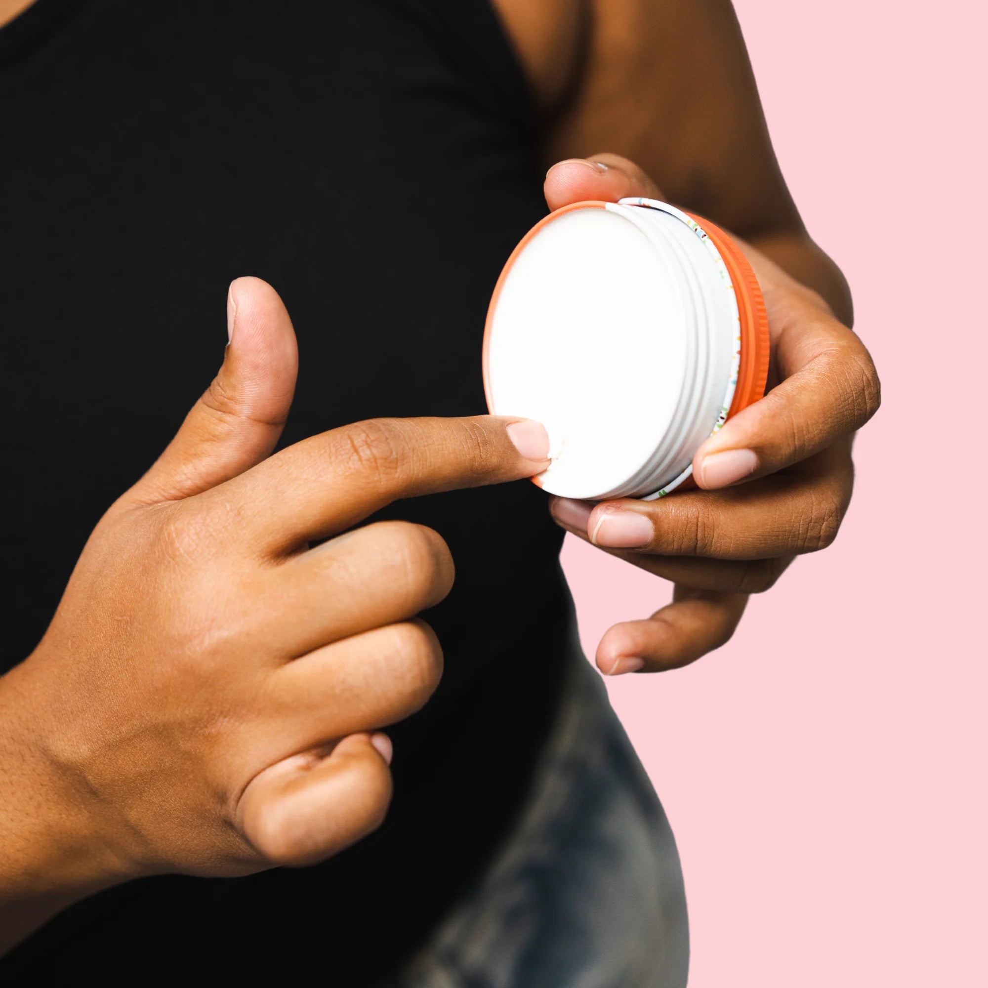 Image of the Woohoo Urban Natural Deodorant Paste tin opened and held by one hand with the finger from the other hand dipping into the product on a pink background