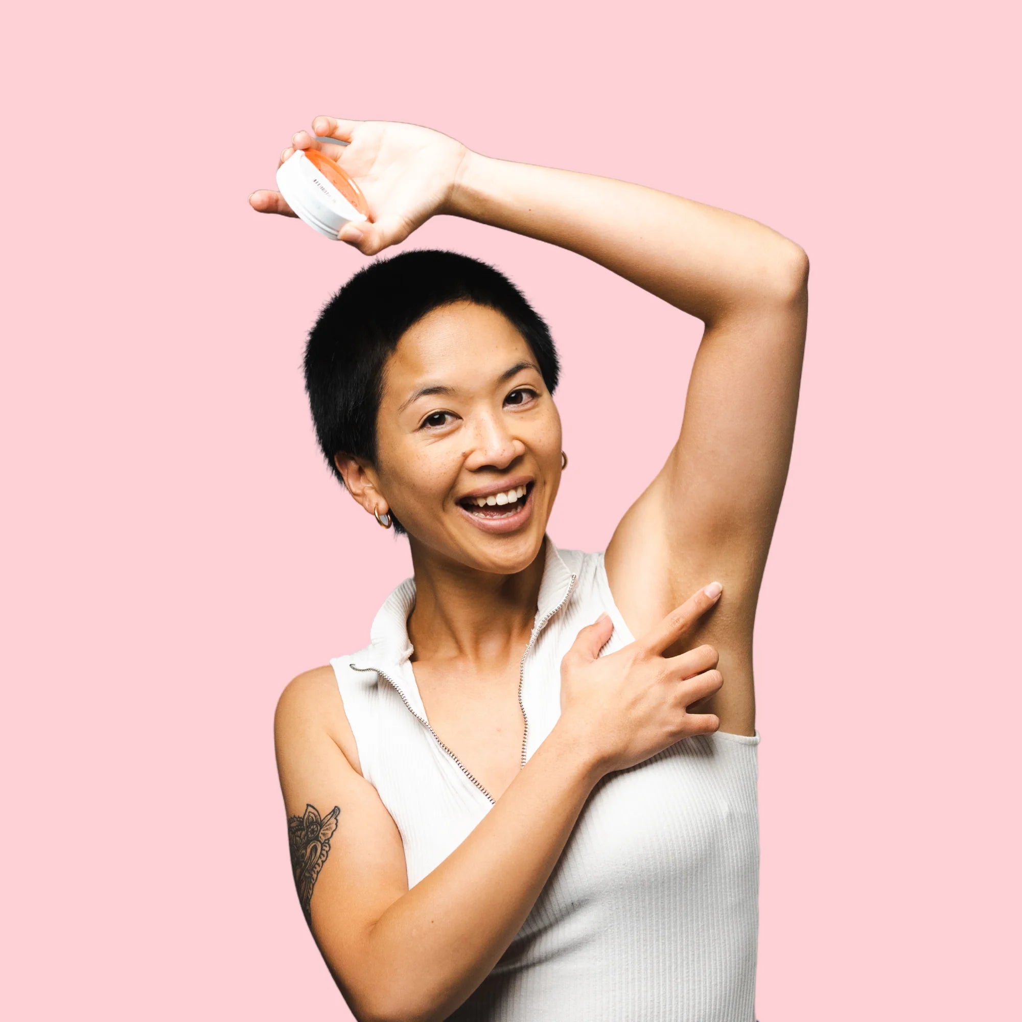 Image of a woman in a white sleeveless shirt holding an Urban Natural Deodorant Paste tin in one hand over her head as she shows her armpit with already absorbed product as she is smiling at the camera in front of a pink background