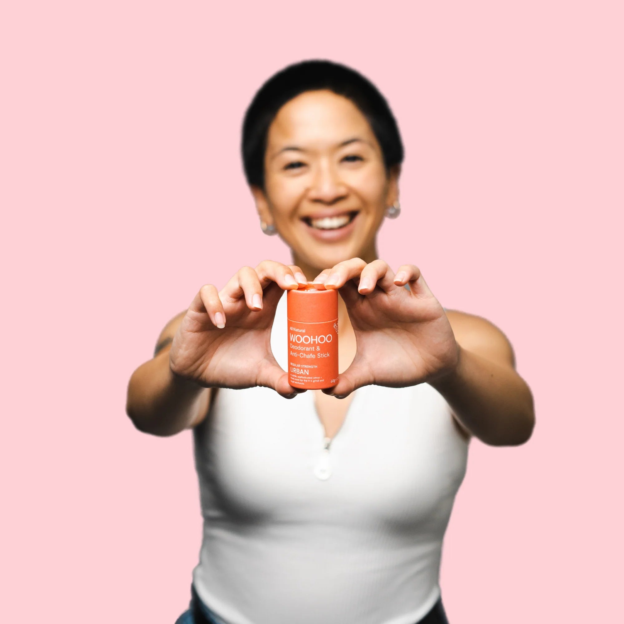 Image of a woman in a white sleeveless shirt smiling to the camera as she holds the Urban Natural Deodorant Stick in both hands extended towards the camera in front of a pink background