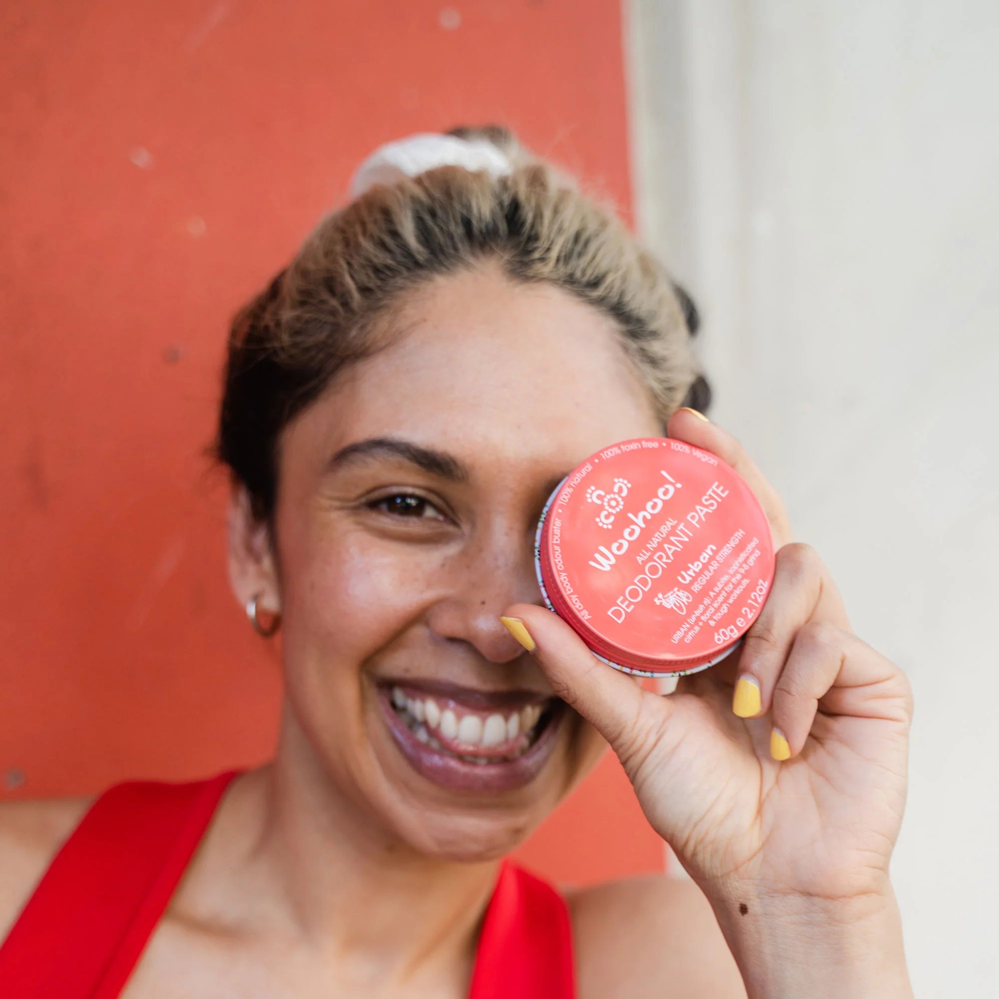 Image of a woman in a red sleeveless shirt holding an Urban Natural Deodorant Paste tin up to her eye as she is smiling at the camera